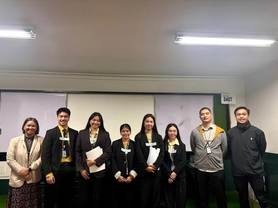 Eight people in dark blazers and jackets standing together in a classroom.