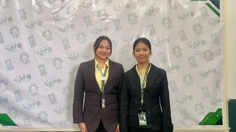 Two students in dark blazers with ID lanyards, standing at a UC InTTO backdrop.